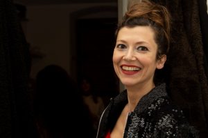 Anajara Amarante, a queer Brazilian woman pictured in a three-quarter view. Smiling she is wearing her brown-redish hair pinned-up, piercings in her ear and nose, black mascara, rose lipstick and a black jacket with a white patterned collar.