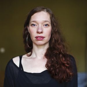 Angela Alves, a white woman with long red wavy hair, looking direcly into the camera. She is wearing a black translucid shirt with a black top underneath as well as large creoles, pink lipstick and black mascara.