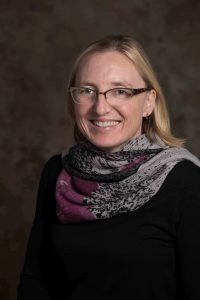 Carrie Sandahl, a white disabled woman with blond hair, sporting glasses and a bright smile, in front of a dark grey background. The photo shows her from the upper body upwards, looking directly at the camera. She is wearing a black sweater and a scarf in purple and grey with black plant ornaments looped several times around her neck.