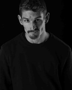 Black-and-white photograph of Dan Daw, a white, disabled, queer man with distinctive dark eyebrows and a beard. He wears a black shirt that is inseparably fused with the background and wrinkles his forehead while direcly looking into the camera.