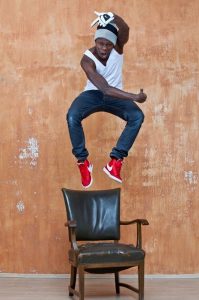 Dodzi Dougban, a black deaf man, jumping high in the air over a dark brown wooden chair with upholstery, in front of a tainted orange wall. He is wearing red Nike shoes, jeans, a white sleeveless shirt, a grey and black beanie and a white Nike glove on his right hand raised over his head.