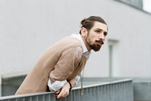 Eyk Kauly, a white deaf man, in three-quarter view looking into the distance as he leans over a balustrade. He wears a brown beard, his long brown hair in a bun, a light brown jumper with a white dress shirt with a collar underneath as well as a long black necklace with a golden key.