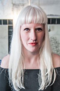 Jo Bannon, a white disabled woman with albinism looking directly into the camera. She is wearing rose lipstick, black mascara and a dark green shirt that leaves her shoulders bare. Her face is framed by her long white hair, which she wears open with a fringe.