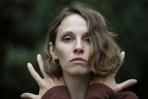 Kassandra Wedel, a white deaf woman, pictured from the shoulders upwards, looking directly into the camera, her light brown, wavy hair falling into her face, partly covering her left eye. She is wearing an auburn long-sleeved shirt and golden creoles. Her hands are raised over her shoulders with splayed fingers, places next to her neck with the outside facing the camera, partly covered from her chin-length hair.