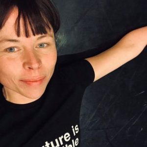 Kate Marsh, a white disabled woman with a red brown fringe lying down on her back on a black dancefloor. Her left arm is spread out in a 90 degree angle from her body. She is wearing a black shirt with the imprint „The future is accessible“ in white letters only partly visible on the photo.