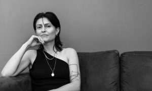 Black-and-white photograph of Rita Mazza, a white deaf Italian woman, sitting on a couch, her head resting on her right hand propped up on the armrest, looking directly into the camera. She is wearing a black shirt that leaves her left shoulder bare, one strand of her dark hair falling into her face between her nose and left eye. She sports painted nails, a tattoo of black lines on her left arm as well as three necklaces, earrings and a ring on her right little finger.